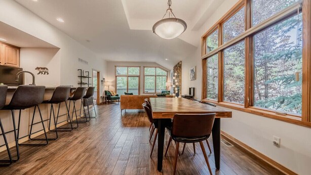 Une cuisine spacieuse avec une longue table en bois et un coin bar, de grandes fenêtres donnant sur la forêt et un luminaire suspendu moderne.