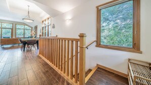 Une pièce spacieuse dotée d’un plancher en bois, d’un coin repas avec une table et des chaises, et de grandes fenêtres offrant une vue sur les arbres.