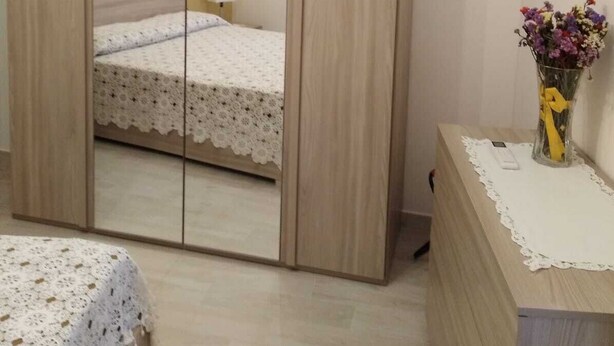 A bedroom with a wooden wardrobe, a bed with a lace bedspread, a bedside table with a vase of flowers, and a nightstand. - Two-roomed house "In centro" + services in the baroque heart of the city (Noto)