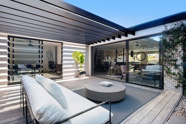 Un área exterior moderna con sala, una mesa redonda y una puerta de vidrio que da a un espacio interior.