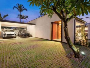Una casa moderna con dos autos estacionados en el garaje, un árbol en el jardín delantero y un camino de ladrillo que lleva a la entrada.