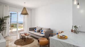 Uma sala de estar moderna com um sofá cinzento, uma área de refeições com mesa de mármore e uma grande janela com vista para o mar.