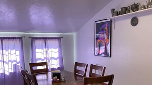 A dining area with a wooden table and chairs, a ceiling light, purple curtains, and a wall-mounted shelf with decorative items.