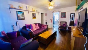 A living room with a sofa, coffee table, and a TV. There are paintings on the walls and a ceiling fan.