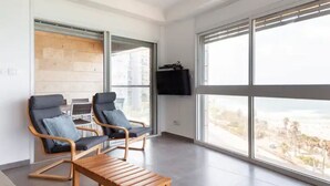 Ein modernes Wohnzimmer mit zwei schwarzen Ledersesseln, einer Holzbank und einem großen Fenster mit Blick auf eine Stadtlandschaft.