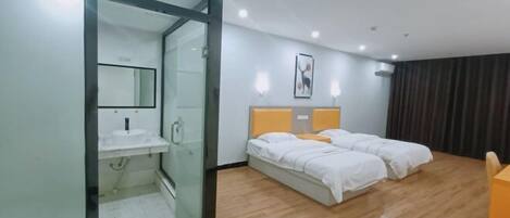 Habitación de hotel con ducha envuelta en vidrio, cama con ropa blanca, mesita de noche y aire acondicionado instalado en la pared.