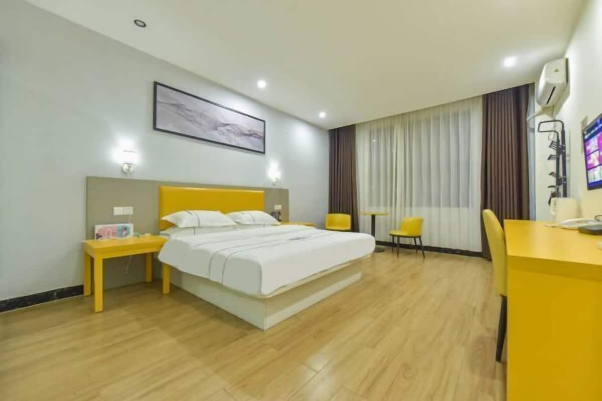 A hotel room with a bed, a desk, two chairs, a bedside table, a wall-mounted TV, and a painting.