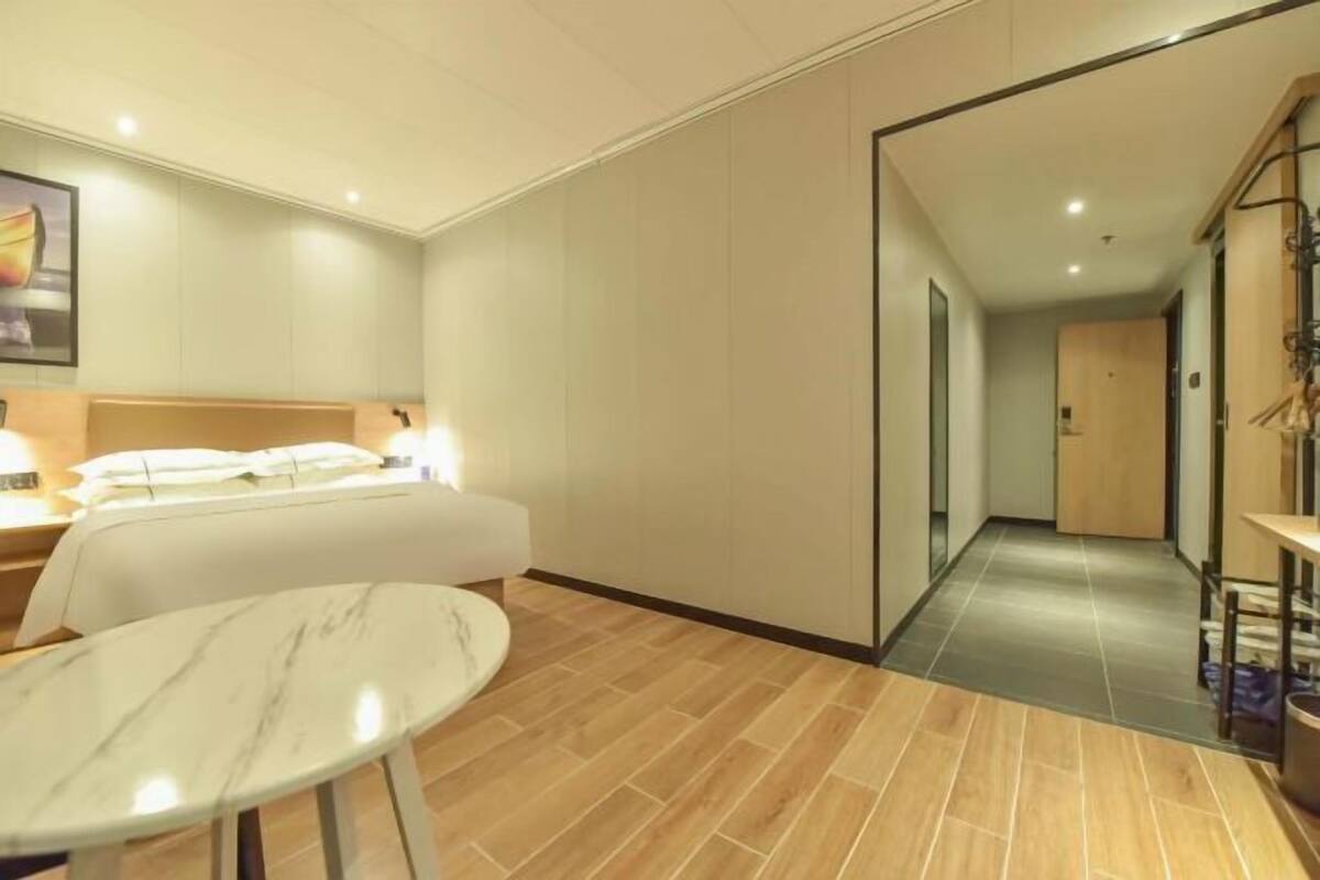 A hotel room with a bed, a bedside table, a chair, a marble table, and a corridor leading to another room.