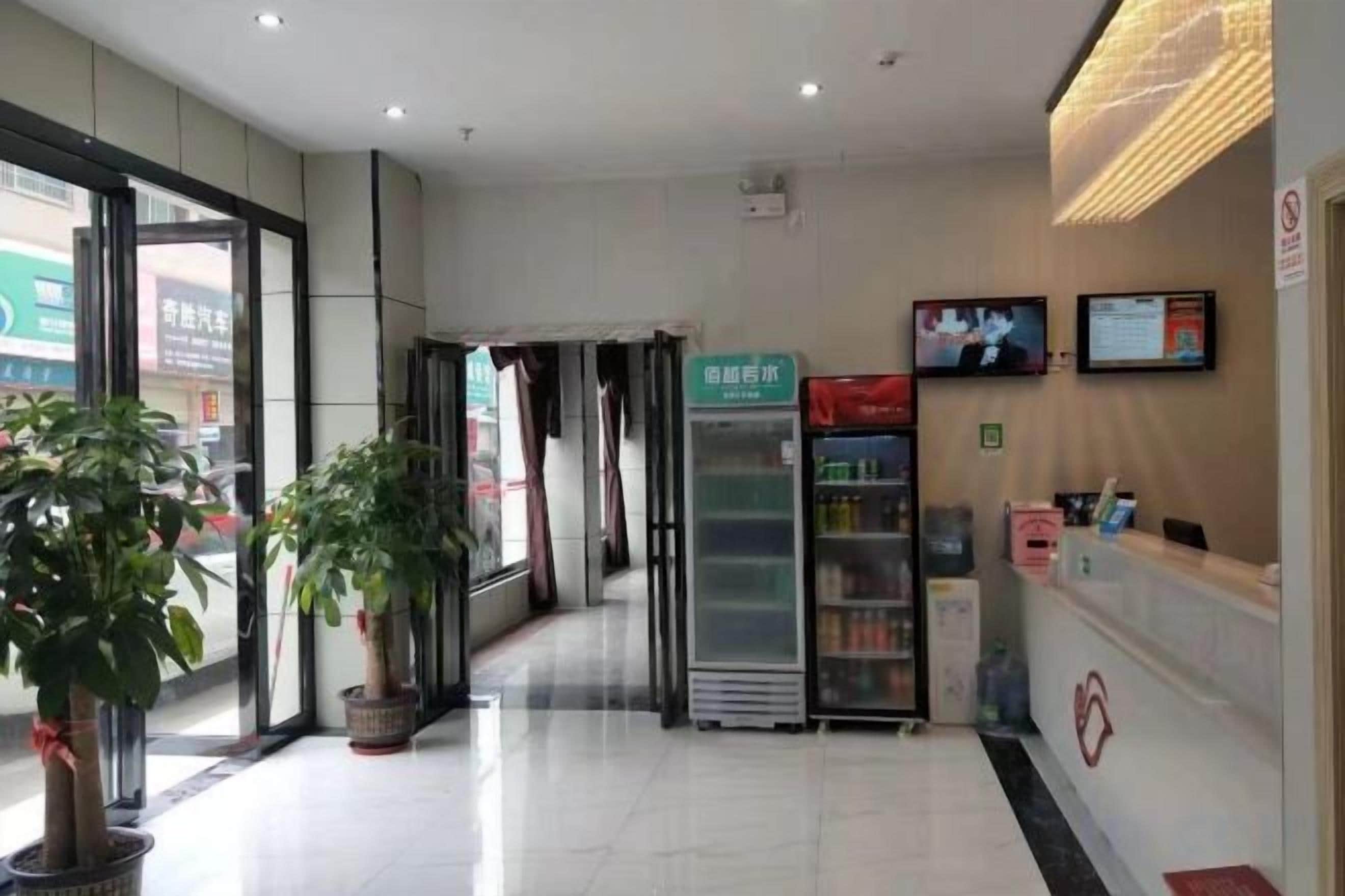 A hotel reception area with a reception desk, a refrigerator, potted plants, and a television screen.