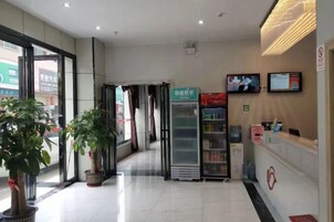Zona de recepción de un hotel con mostrador, refrigerador, plantas en macetas y una pantalla de televisión.