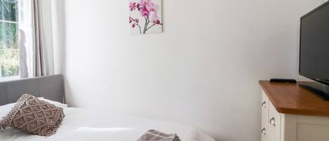 Una cama bien hecha con una toalla doblada, un cojín decorativo, un cuadro de flores en la pared y un televisor sobre un aparador de madera.