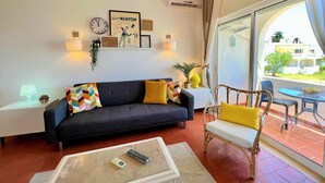 Ein Wohnzimmer mit einer Couch, Sesseln, einem Couchtisch und einem Fernseher. An den Wänden sind Lampen befestigt, eine Uhr ist zu sehen, und es gibt gerahmte Kunstwerke.