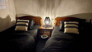 A room with two single beds, a small table with a lamp, and a waste bin.