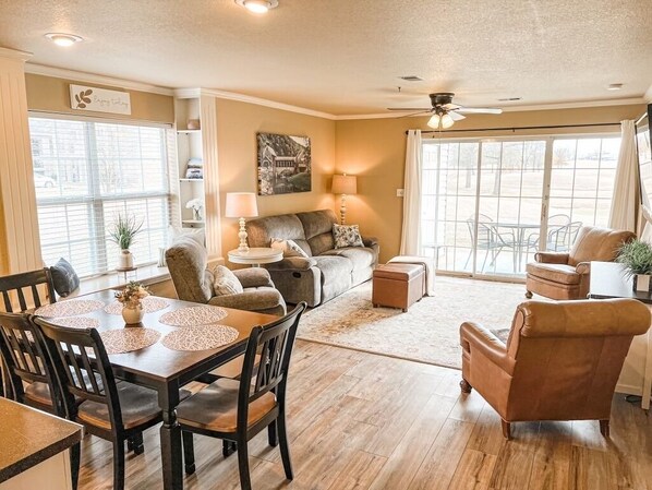 A spacious living room with a sofa, a dining table with chairs, a ceiling fan, and large windows.
