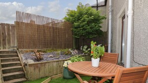 Une terrasse en bois avec une table et des chaises, entourée d’un jardin agrémenté d’une clôture en bois, d’un petit étang et de diverses plantes.