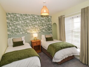 A bedroom with two beds, a nightstand with a lamp, a wall with a tropical leaf pattern, and a window with curtains. - Greystones Cottage (Crieff)