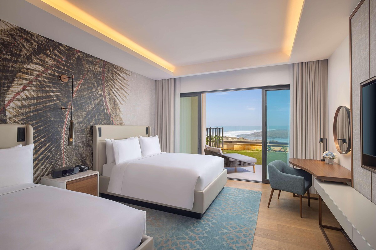 A hotel room with a large bed, a desk, a chair, a TV, and a balcony with a view.