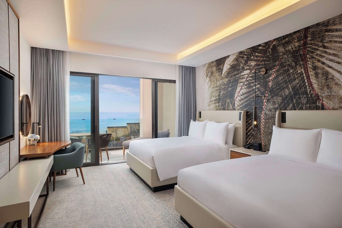 A hotel room with two beds, a desk, a chair, a TV, and a large window with a view of the sea.