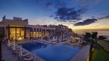A modern hotel with a pool, sun loungers, and a view of the sunset over the ocean.