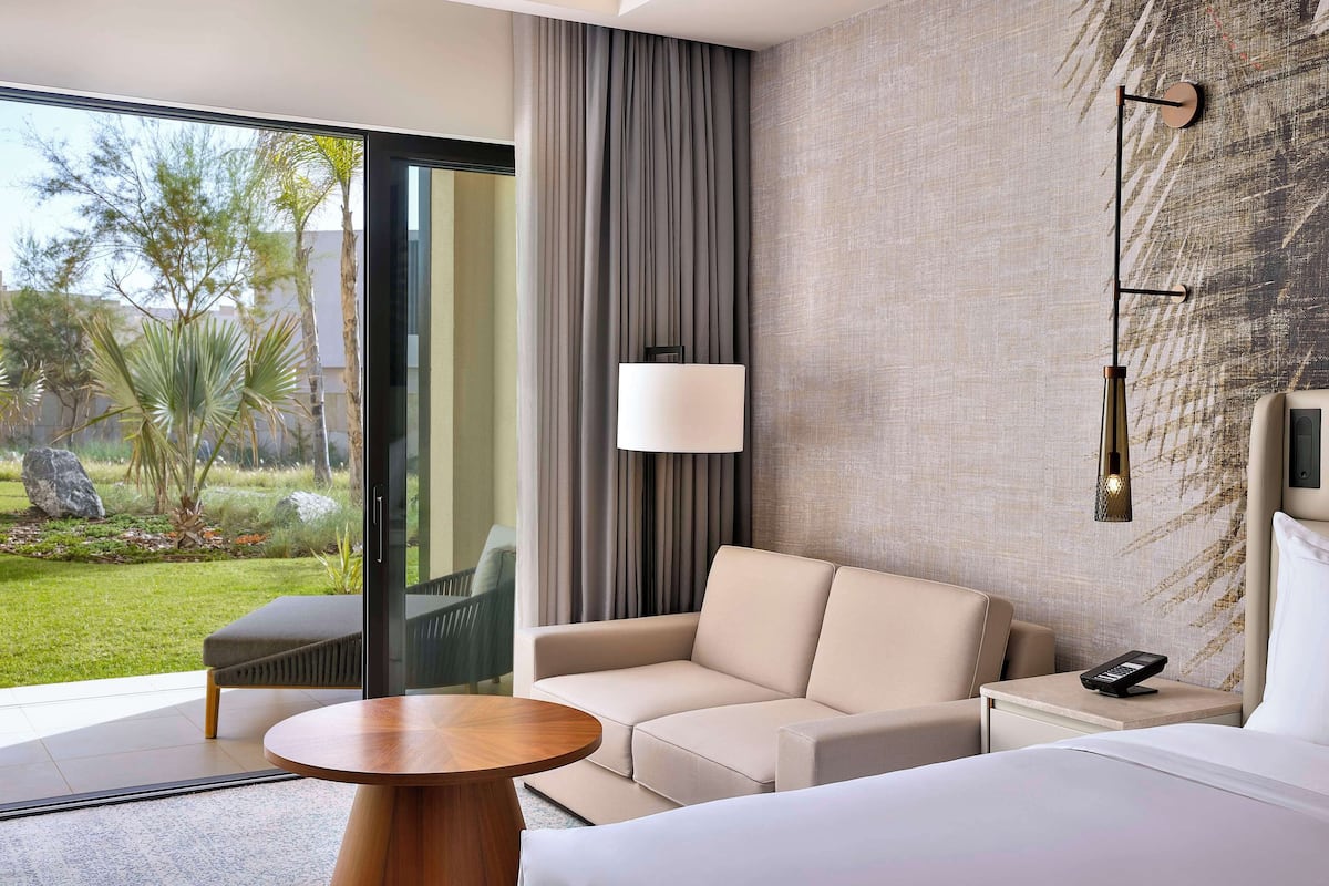 A modern hotel room with a sofa, a small table, a bedside table with a lamp, and a view of a garden through large windows.