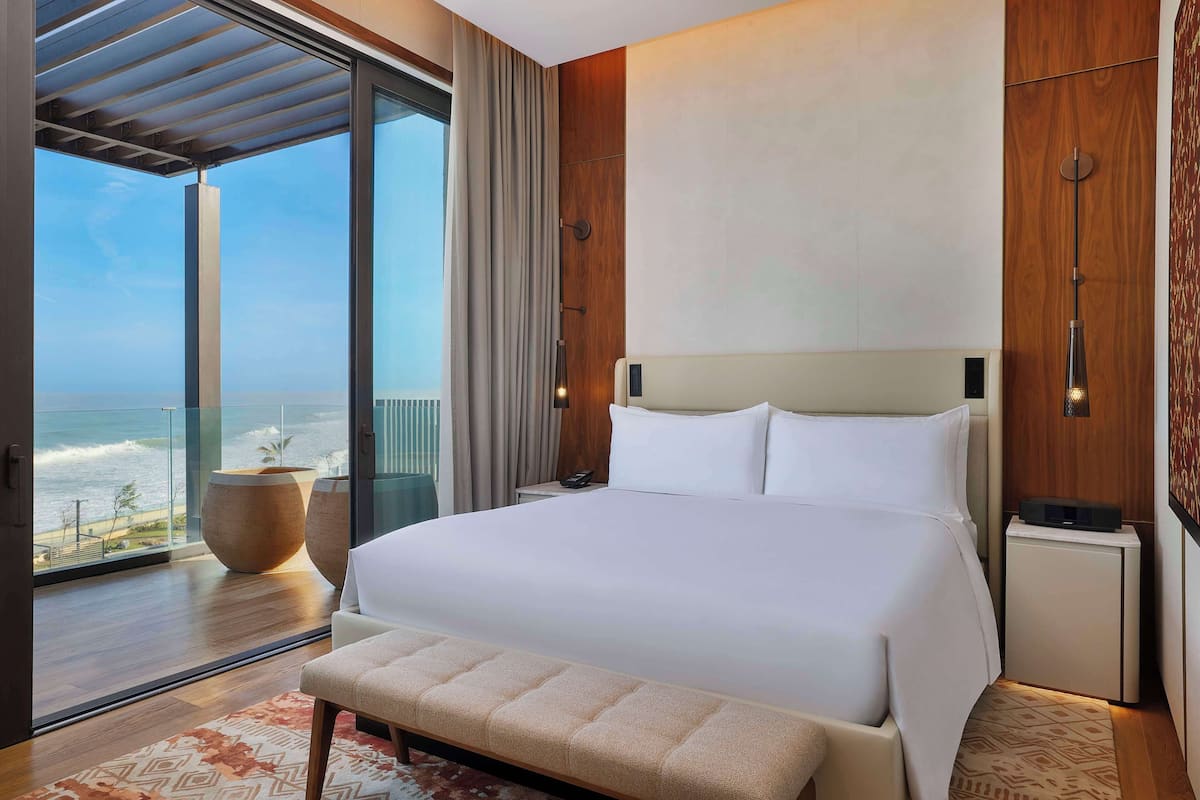 A modern hotel room with a large bed, a bench, a nightstand, and a view of the ocean.
