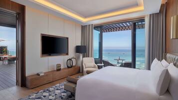 A modern hotel room with a large bed, a TV mounted on the wall, a seating area with a view of the ocean, and a balcony with a table and chairs.