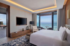 A modern hotel room with a large bed, a TV mounted on the wall, a seating area with a view of the ocean, and a balcony with a table and chairs.