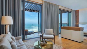 A modern hotel room with a sofa, armchair, and a coffee table. There is a view of the ocean through large windows. - Conrad Rabat Arzana (Harhoura)