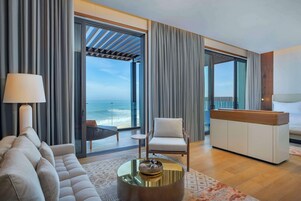 Quarto de hotel moderno com sofá, poltrona e mesa de centro. Há vista para o oceano através de grandes janelas.