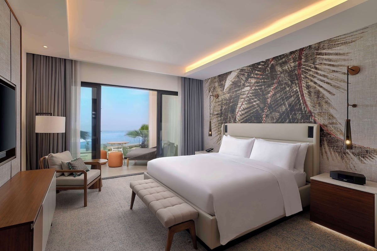 A modern hotel room with a large bed, a desk, a bench, and a view of the ocean.