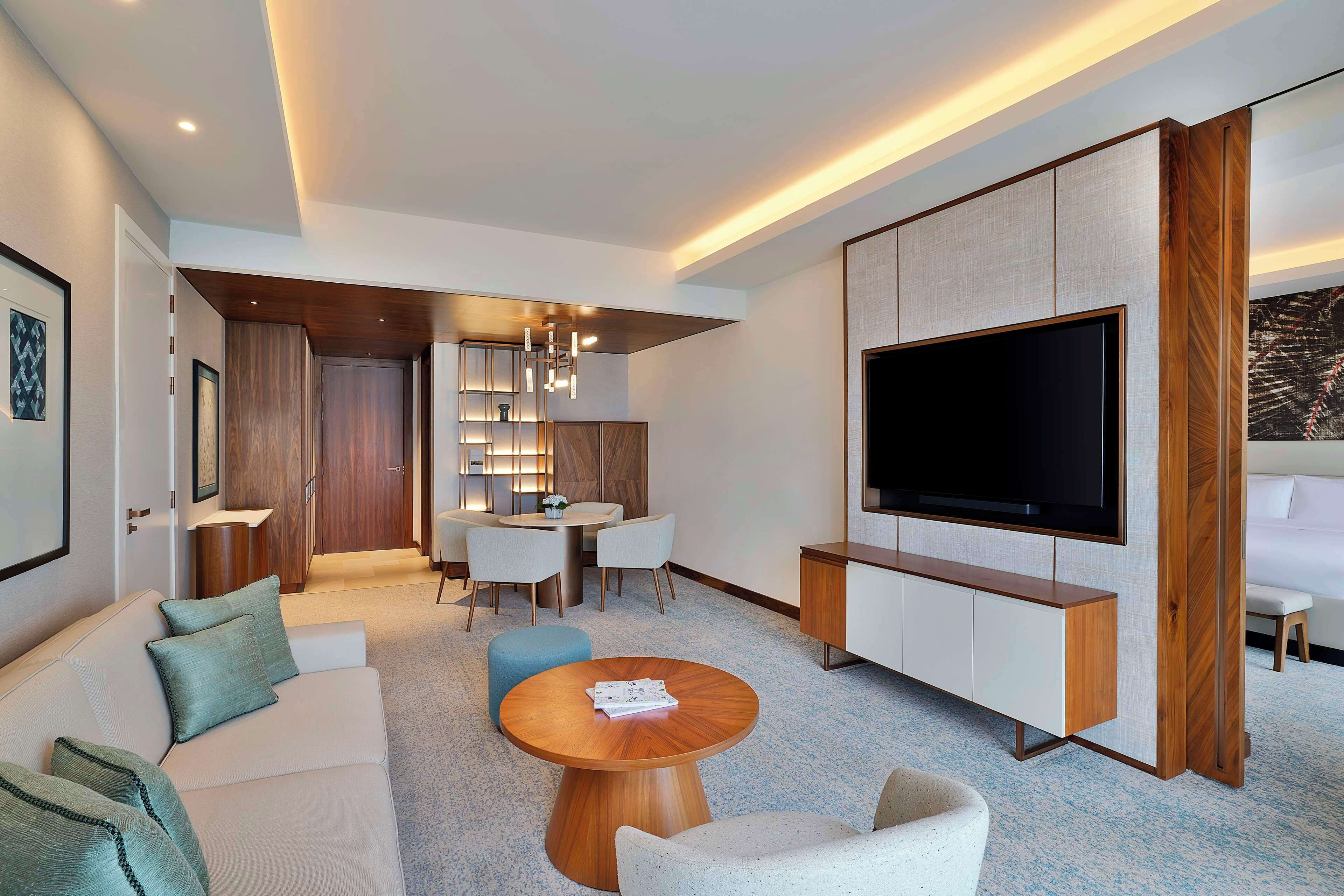 A modern hotel room with a sofa, a round wooden coffee table, a TV mounted on the wall, and a dining area with a table and chairs.