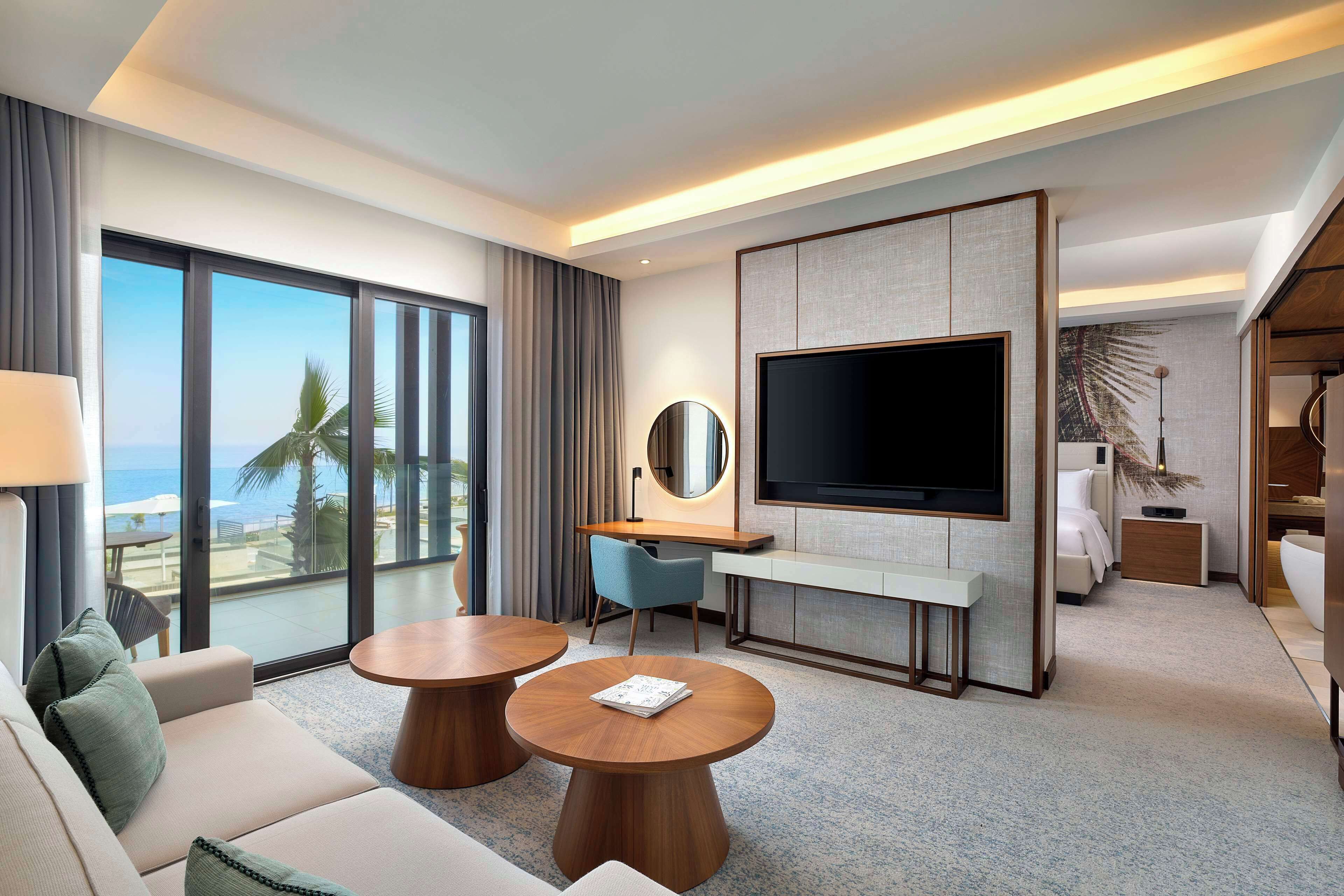 A modern hotel room with a large TV, a sofa, two round tables, a desk, and a view of the ocean.