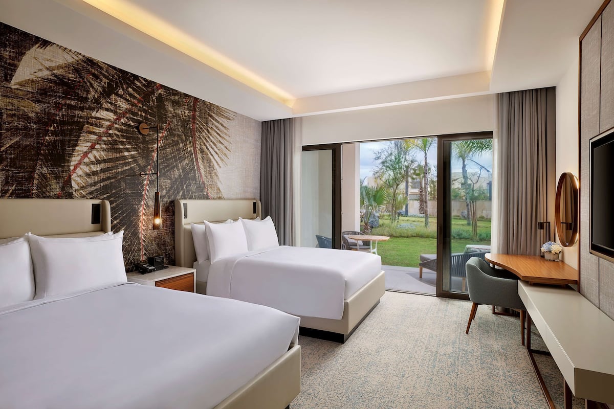A hotel room with two beds, a desk, a chair, a TV, and a large window with a view of the outdoors.