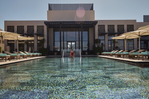 A swimming pool with lounge chairs and umbrellas, a modern building with large windows, and a clear sky.