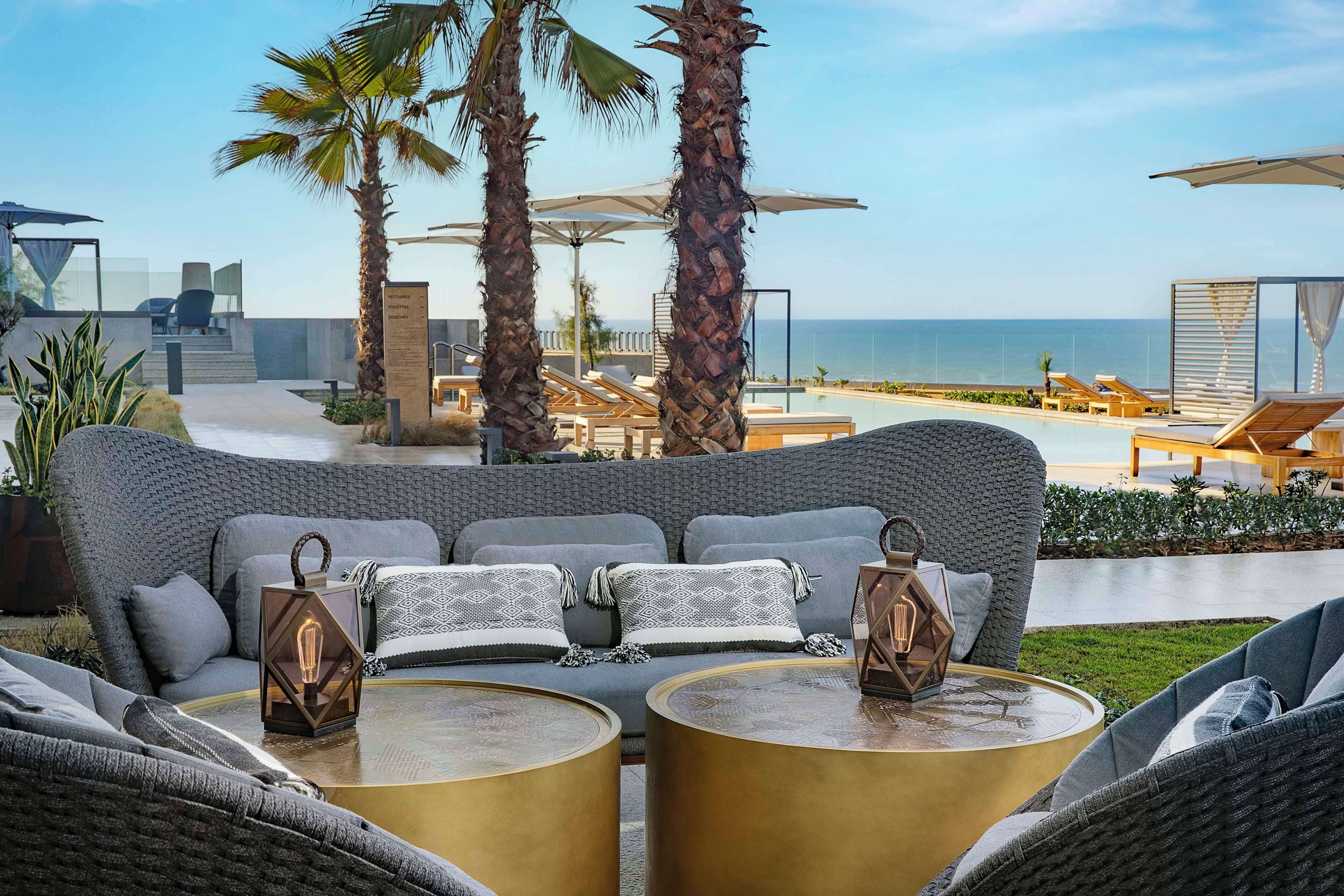 Een moderne buitenlounge met een bank en twee ronde tafels, met uitzicht op een zwembad en de oceaan.