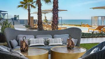 A modern outdoor lounge area with a sofa and two round tables, overlooking a pool and the ocean.