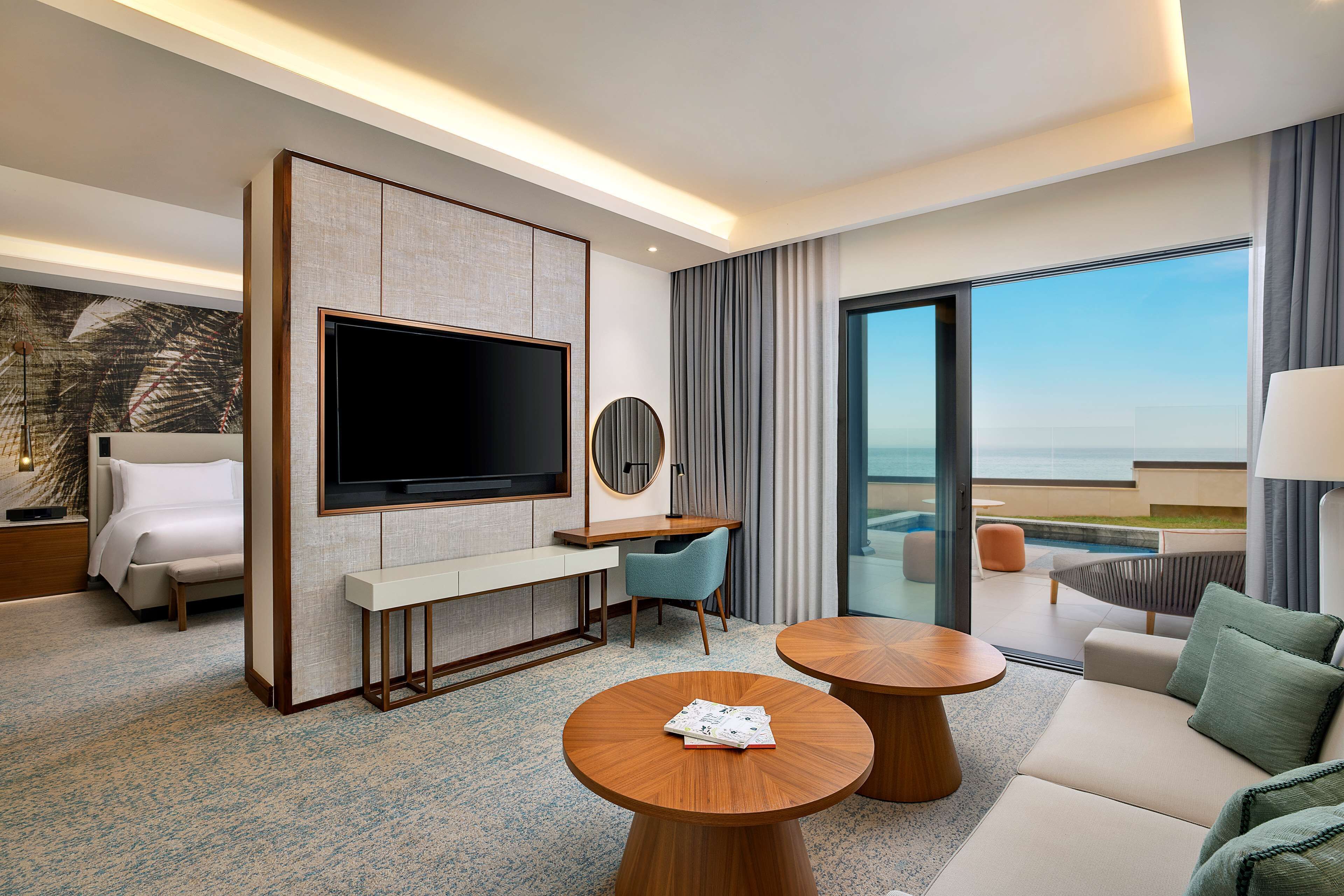 A modern hotel room with a large TV, a desk, a sofa, and a balcony with a pool view.