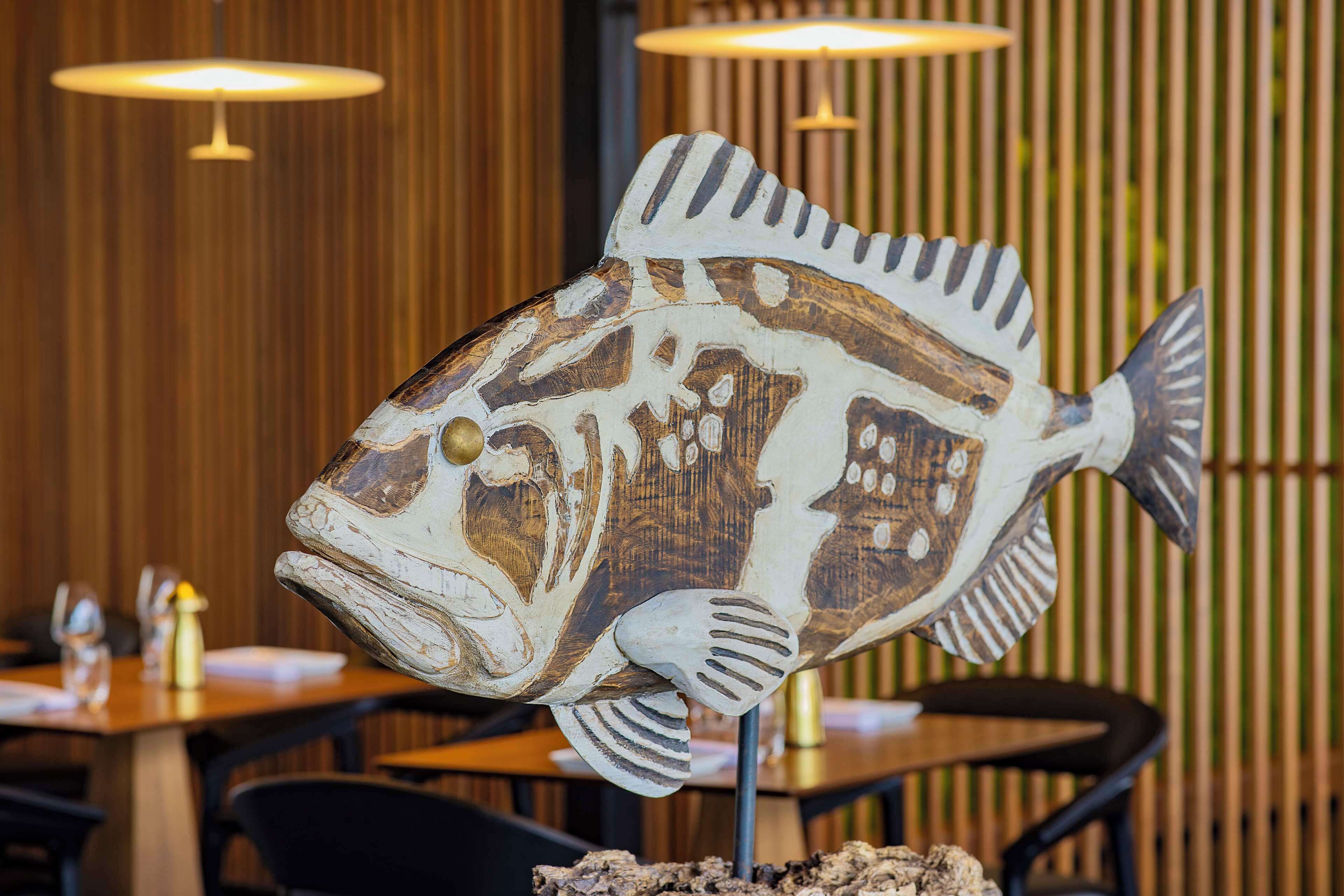 A wooden fish sculpture on a table in a restaurant with vertical wooden slats in the background.
