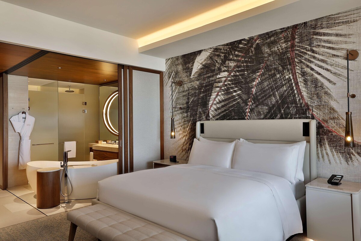 A modern hotel room with a large bed, a bathroom with a bathtub, and a wall with a leaf pattern.