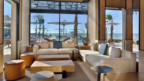 A modern lounge area with a sofa, ottomans, and a coffee table, overlooking a pool and palm trees. - Conrad Rabat Arzana (Harhoura)