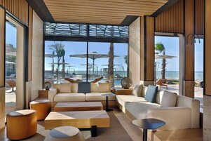 Uma área de estar moderna com um sofá, pufes e uma mesa de centro, com vista para uma piscina e palmeiras.