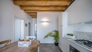 A modern kitchen with wooden beams, white cabinetry, a gas stove, and a dining area with a table set for two and a fruit bowl. - Modern beach apartment with parking space (Chioggia)