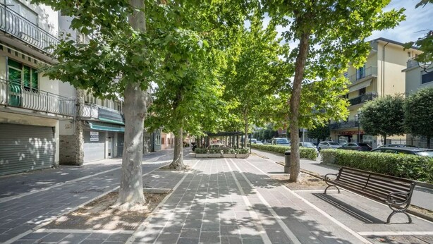 A paved pedestrian walkway lined with trees and benches, with buildings on either side. - Modern beach apartment with parking space (Chioggia)