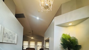 Um corredor com um candelabro, uma placa de boas-vindas, uma planta em vaso e um espaço de estar com sofá e poltronas.