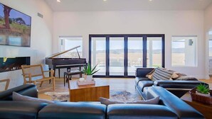 A spacious living room with a grand piano, comfortable seating, and large windows offering a view of the outdoors.