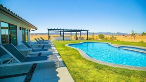 A swimming pool with lounge chairs and a pergola, surrounded by a well-maintained lawn and distant mountains.
