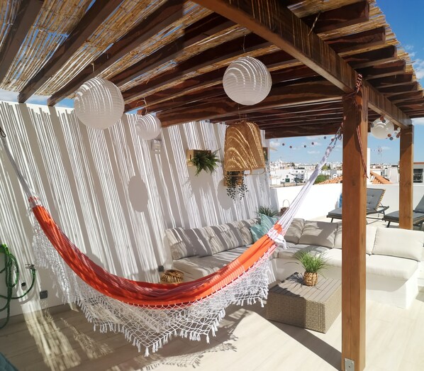 A hammock under a wooden pergola with a white wall and a view of the city.