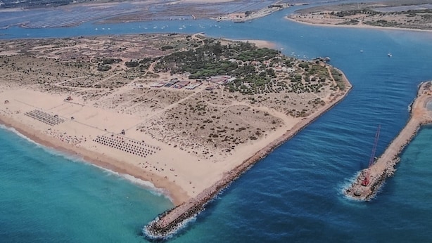 Vue aérienne d’une zone côtière avec une plage de sable, une petite île et une jetée qui s’avance dans l’eau.