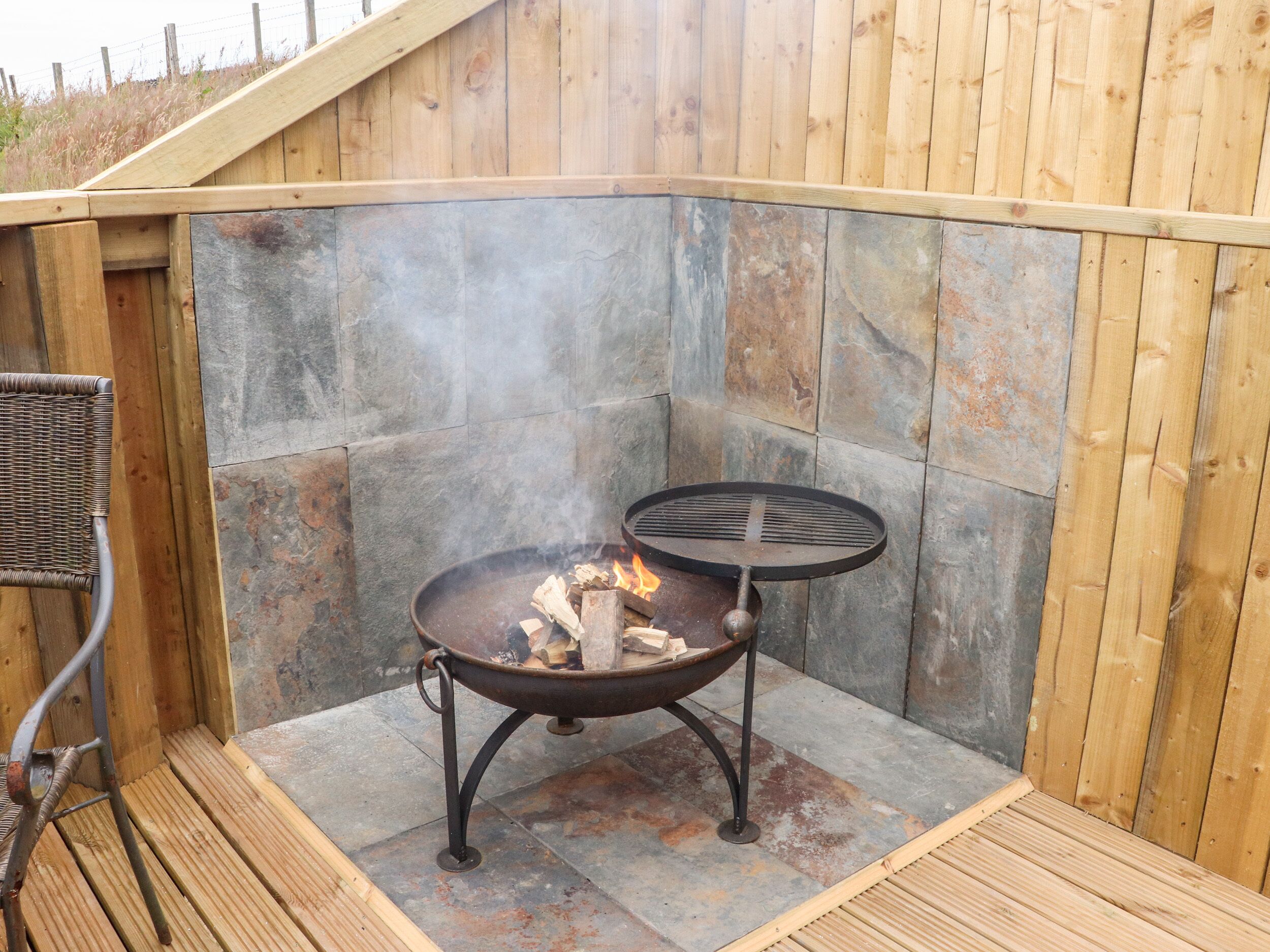 A stone fire pit with a metal table, surrounded by wooden decking and a wicker chair.