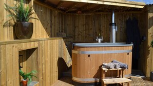 Een houten kuub met een hottub, een bank en handdoeken, omringd door houten wanden en potplanten.